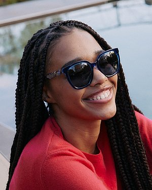 Frau lächelt im Freien und trägt eine blaue Esprit-Sonnenbrille mit gemusterten Bügeln und einen roten Pullover.