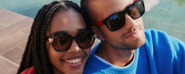 Zwei Models, die Esprit-Sonnenbrillen mit gewagten, farbigen Akzenten tragen, lächeln und sich draußen bei einem Pool befinden.